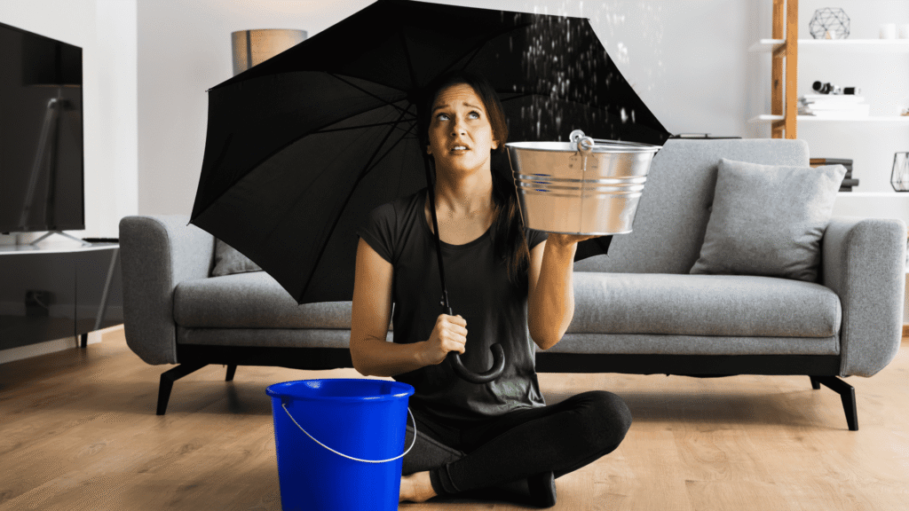 Woman sitting in a living room holding an umbrella and a bucket due to a ceiling leak, requiring immediate restoration service in Kissimmee.
